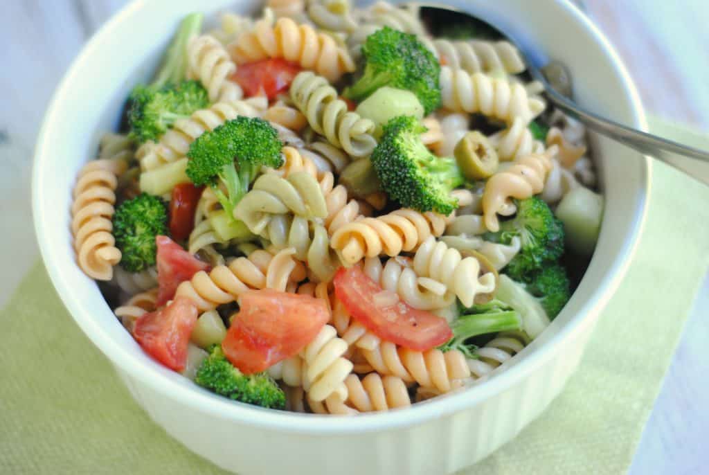 white bowl on a green napkin background filled with an Italian Broccoli and Pasta Salad of tri color pasta mixed with broccoli, tomatoes, and peppers. There is a spoon on the right.