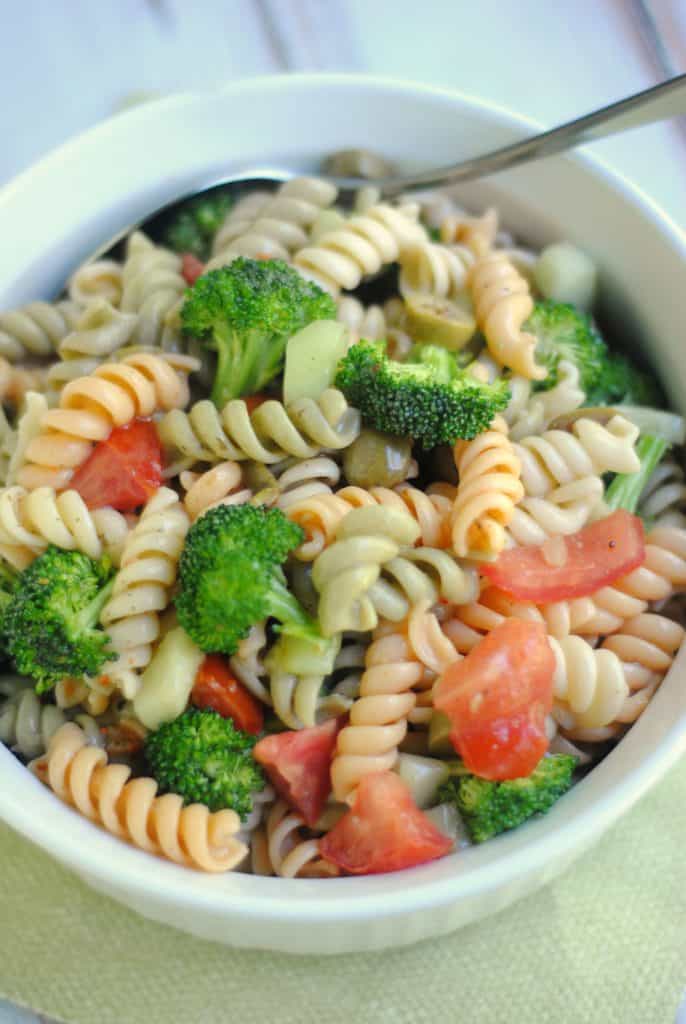 white bowl on a green napkin background filled with an Italian Broccoli and Pasta Salad of tri color pasta mixed with broccoli, tomatoes, and peppers. There is a spoon on the right.