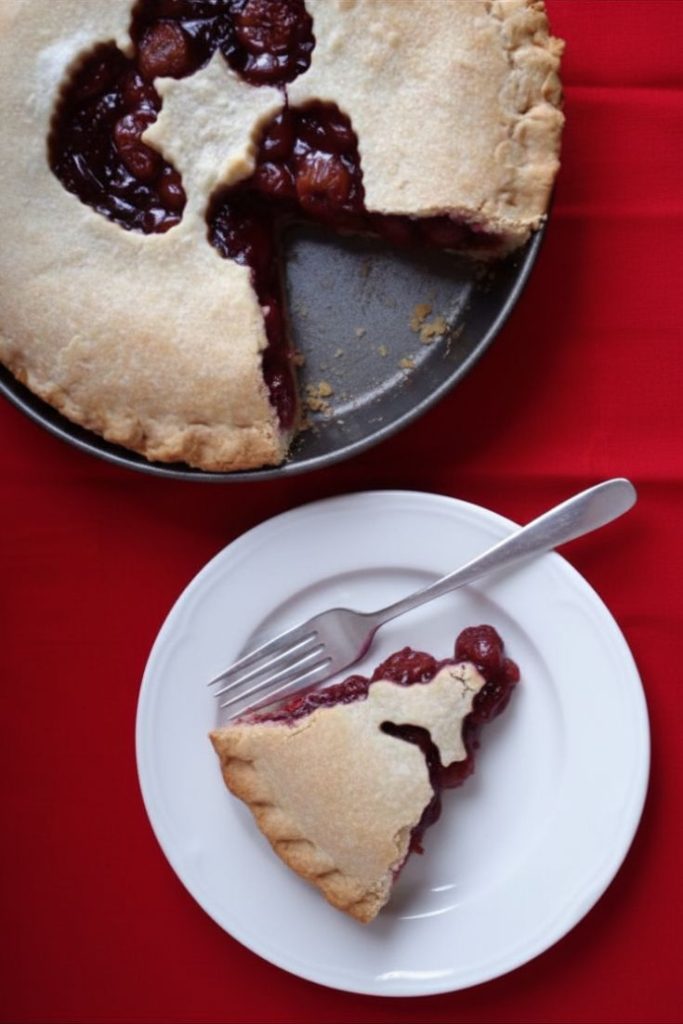 Cherry pie in a pan with a slice cut out of it.  The slice is on a white plate with a fork next to it.