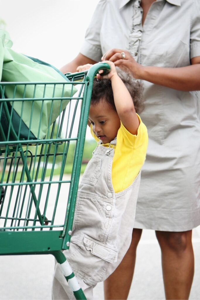 A 2 year old hanging on the back of a shopping cart with a mom standing behind them.